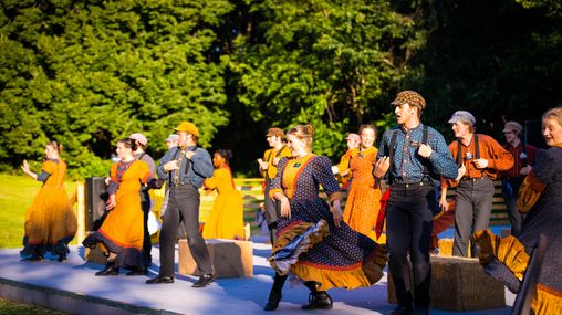 Missionary performers dancing on an outdoor stage dressed in pioneer era clothing. 