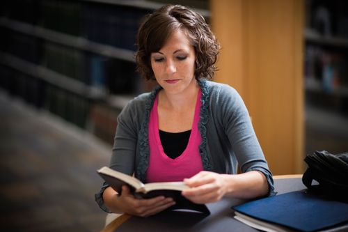 A young woman with curly, short brown hair sits at a round table in a library and reads from a book, with bookshelves in the background.