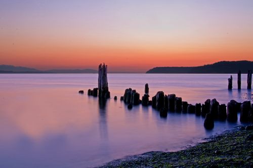 An old pier extends out into the water where purple light is reflected after the sun sets on the horizon.