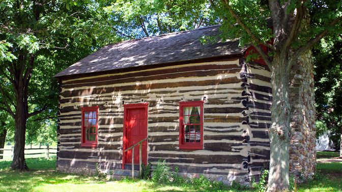 A log cabin with a red door and red-framed windows, surrounded by trees in Fayette, New York.