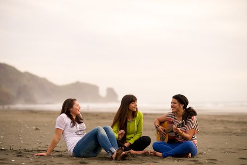 A young woman plays her guitar on the beach while two other young women sit on the sand with her, with water and mountains in the background.