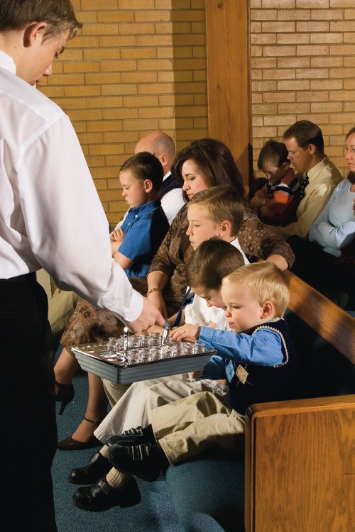 A small boy with blonde hair reaches out to take a cup from a metal sacrament tray that a deacon is holding out to him.