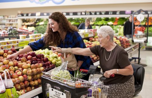 young adult woman helping older woman in a motorized chair reach some apples at grocery store