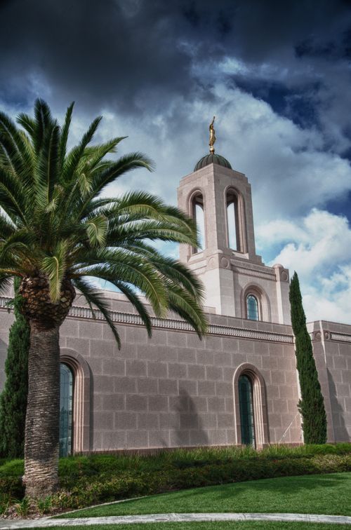 The Newport Beach California Temple and spire, with darkening clouds overhead and a palm tree growing near the temple.