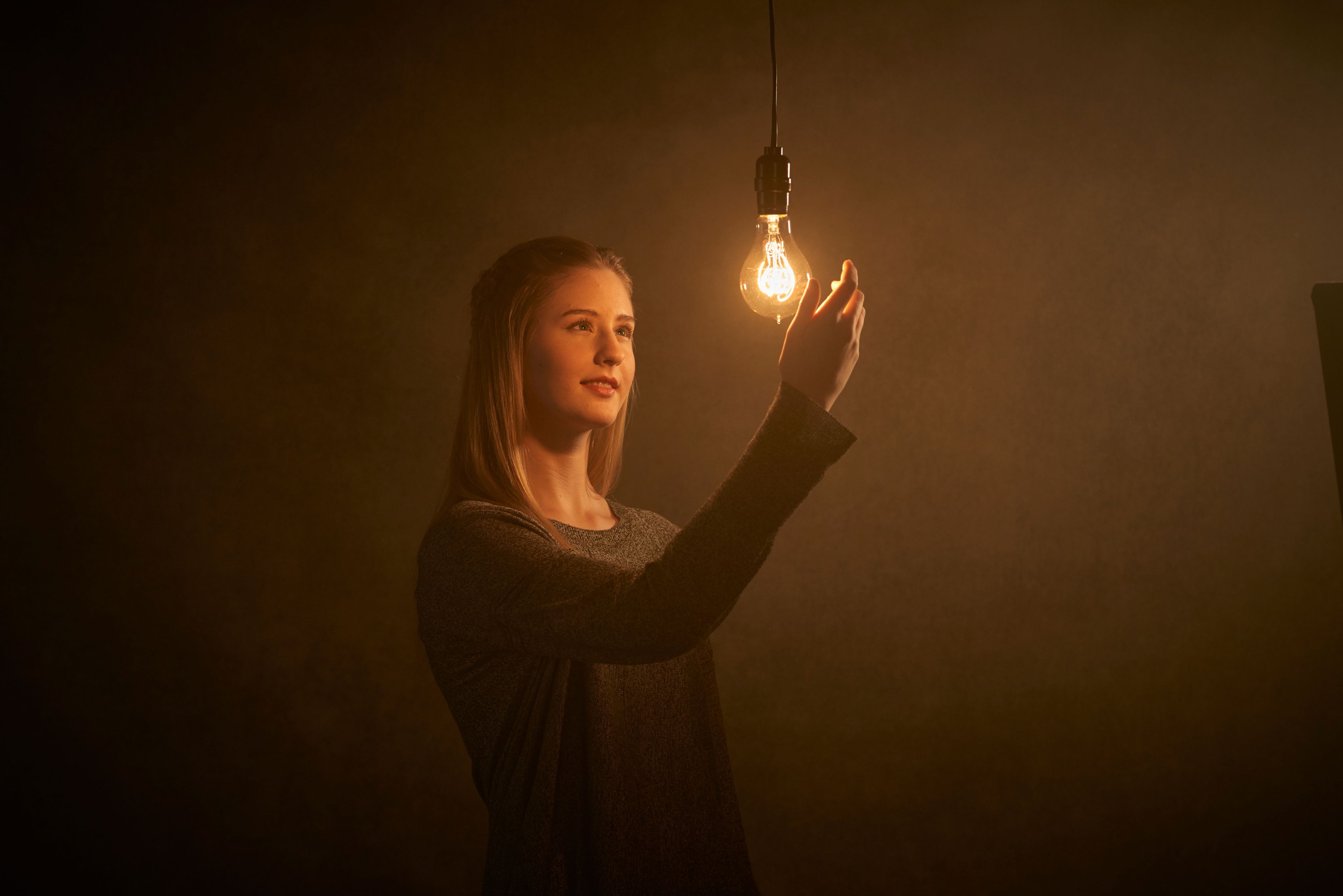 A young woman in a dark room reaching out toward an illuminated light bulb.