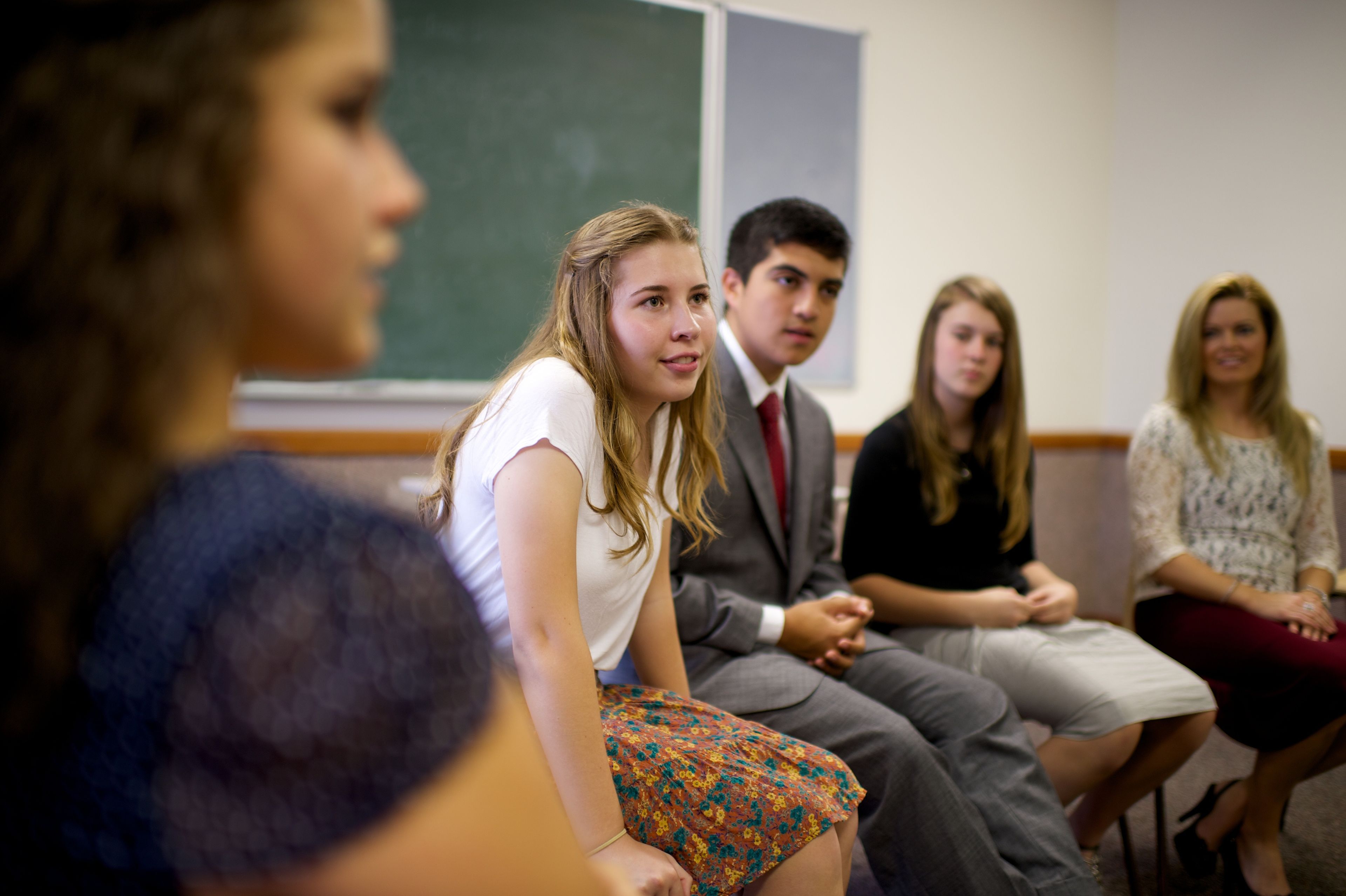 A young woman leans forward in her chair and listens to a teacher in Sunday School surrounded by youth.