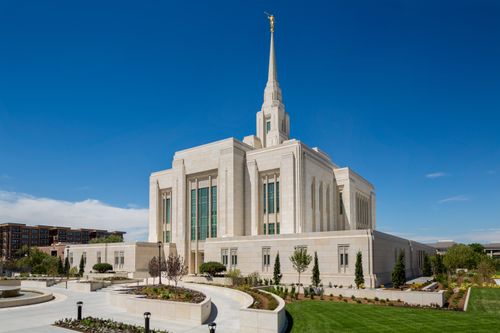 A front side view of the Ogden Utah Temple, with green lawns and some of the surrounding buildings on the street in the frame.