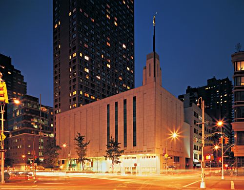 A view of the Manhattan New York Temple front entrance in the late evening, lit up by surrounding street lights, with buildings seen in the distance.