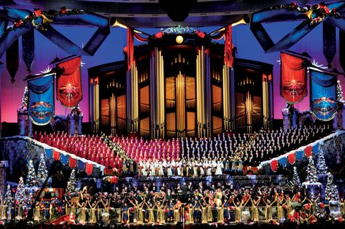 The Mormon Tabernacle Choir and Orchestra at Temple Square onstage at the Conference Center, which is decorated with Christmas trees and banners.