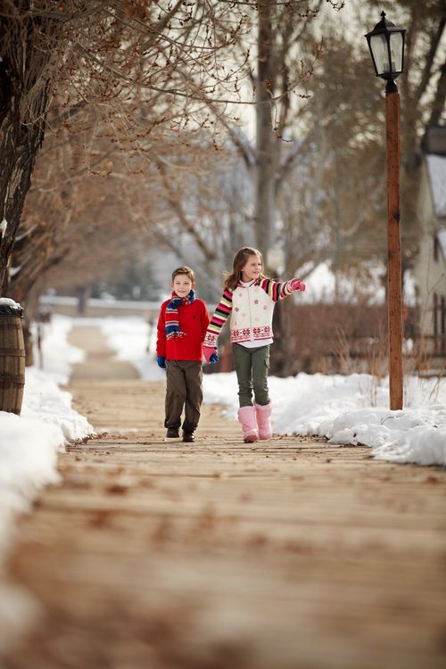 A brother and sister hold hands while walking down a sidewalk, with snow on either side.