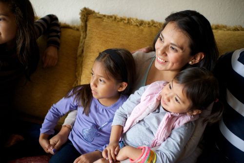 A dark-haired woman sits on a yellow couch, holding her two young daughters, who are posing for another camera that is out of the frame.