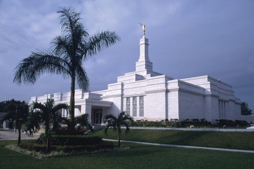 The Mérida Mexico Temple viewed at an angle, showing the side and front entrance, with some of the gardens of flowers and palm trees on an overcast day.