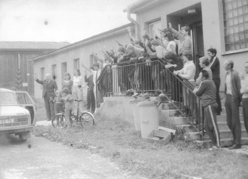 About fifty people stand on the steps of a building and wave to a car.