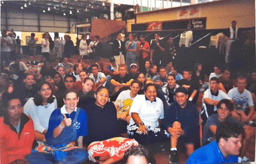 Sixty youth sit on the floor of a gymnasium and smile at the camera.