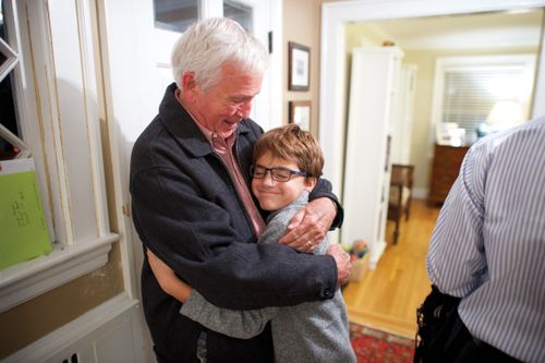 An elderly man gives his grandson a hug in the entryway of the house.