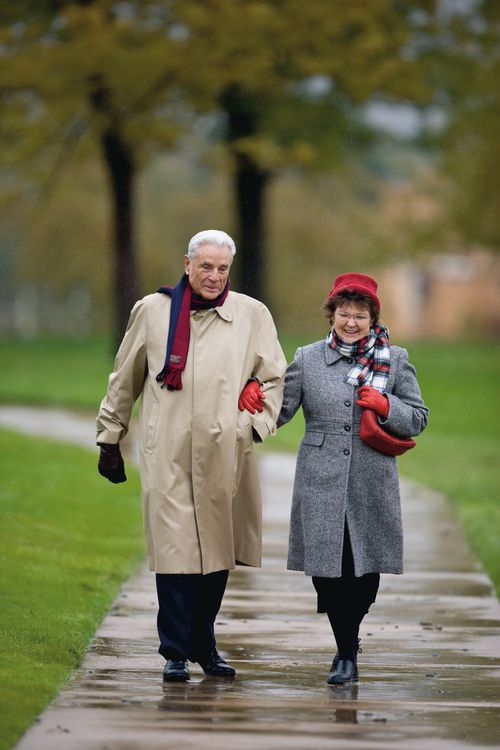 An elderly couple in coats, scarves, and gloves, walking with arms linked down a wet sidewalk.