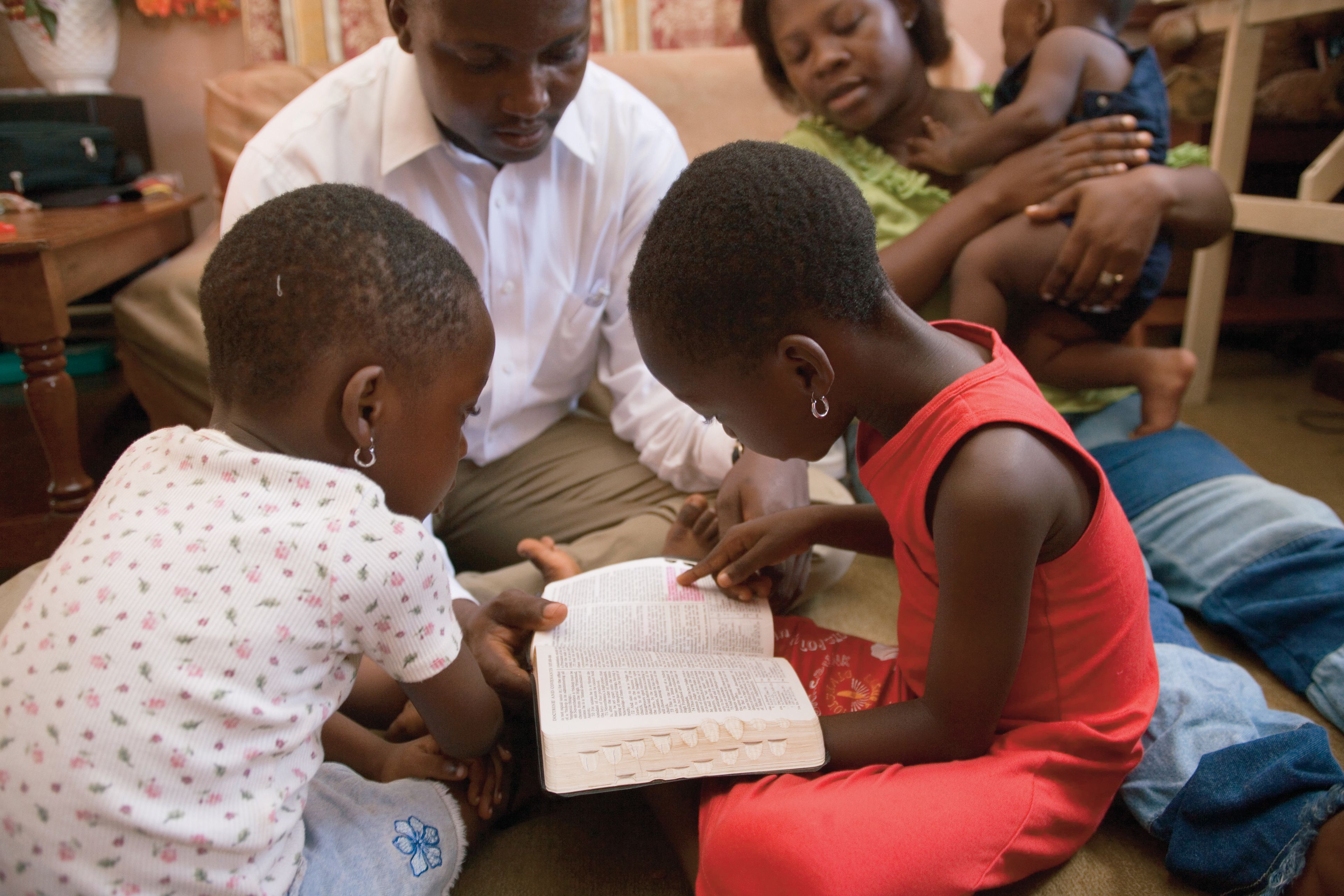A father helps his two daughters read from the scriptures.