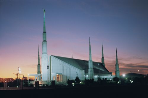 A side view of the Buenos Aires Argentina Temple lit up, with rows of lights on the ground at dusk.