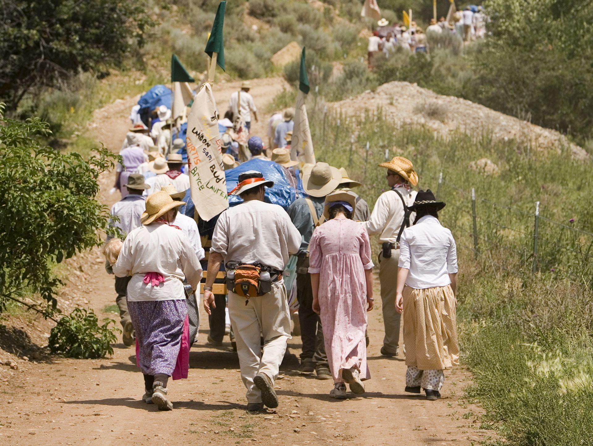 A group of men and women, all wearing hats, walk on a dirt road up a hill and pull handcarts.