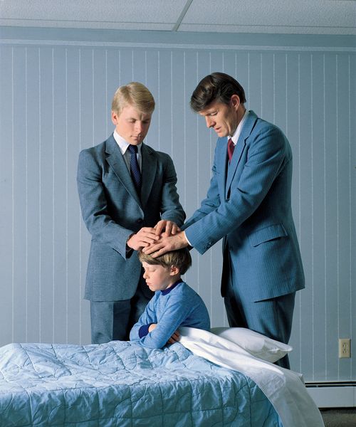 A young boy in blue pajamas sitting up in his bed while two men in suits lay their hands on his head to give him a blessing.