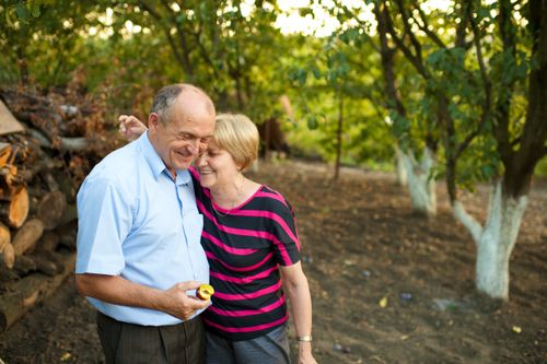 An elderly woman standing with her arm over her husband’s shoulder while he holds a piece of sliced fruit in an orchard in Romania.