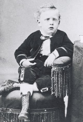 George Albert Smith around four years old, wearing a jacket, pants, and lace-up boots, sitting in a chair and leaning his arm on the armrest.