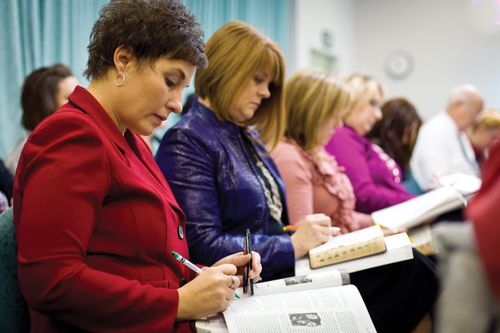 A woman in a red blazer sits on a chair with a manual and a set of scriptures open on her lap, next to several other women in institute.