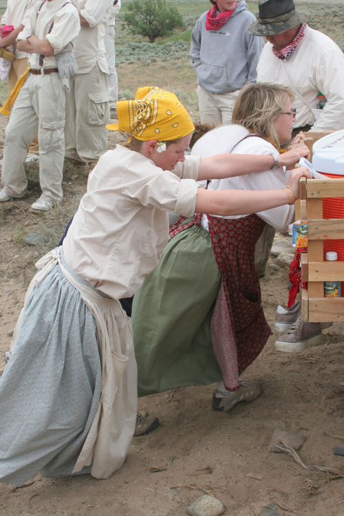 Two women in skirts and white shirts push a handcart uphill over rocks and a dirt path.