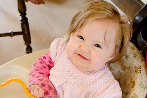 A baby girl sitting in a high chair, looking up and smiling.