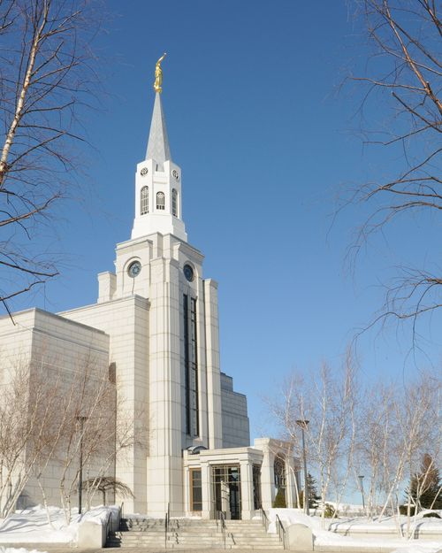 The entrance and spire of the Boston Massachusetts Temple in the winter, with bare trees and snow on the grounds.