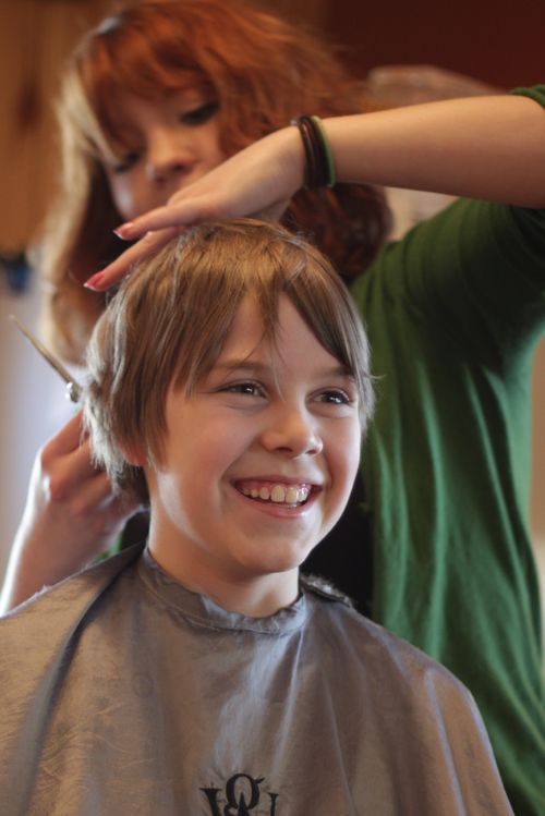 A young boy smiles while a woman stands behind him and cuts the back of his hair.
