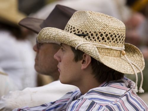 A young man with dark brown hair, wearing a striped button-up shirt and straw hat, sits down with a group of people during trek.