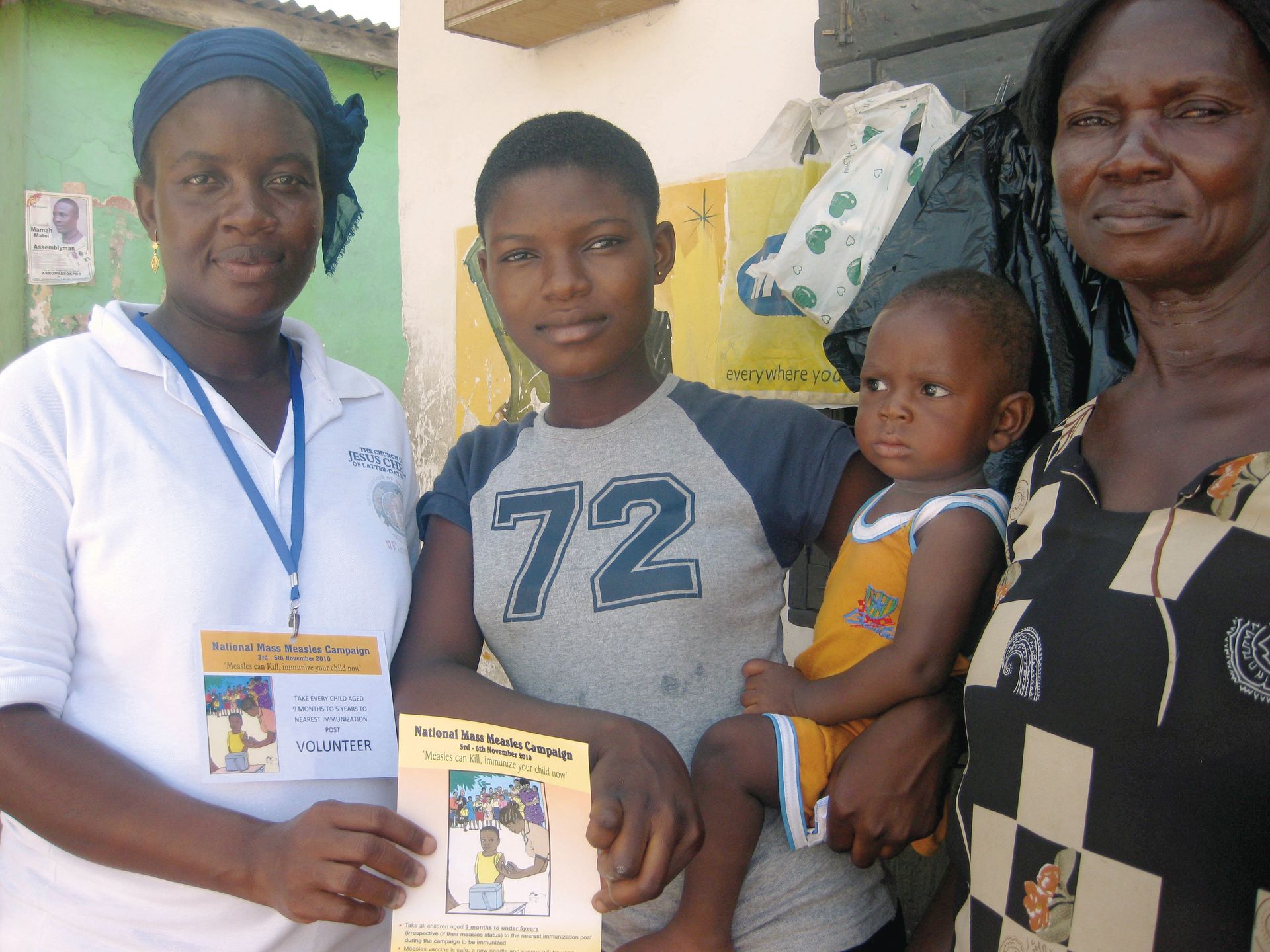 A woman in Africa holding a measles campaign flier and standing near another woman, young woman, and child.