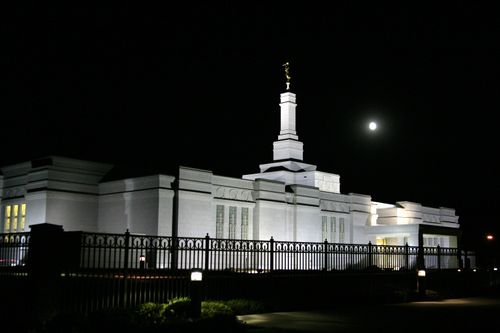 The side of the Spokane Washington Temple, with a view of the fence around the grounds, all lit up at night.
