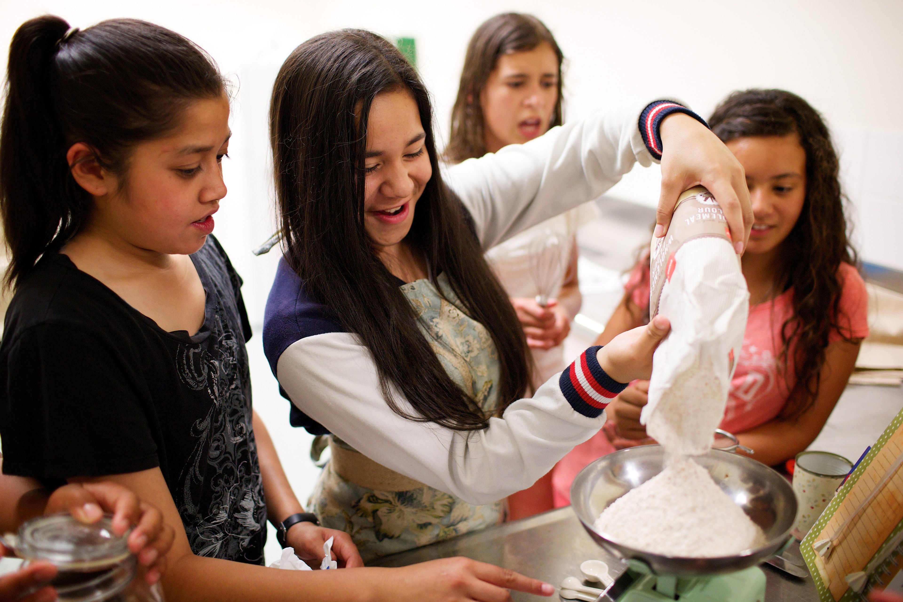 A young woman pours flour into a bowl as she and three other young women bake together.