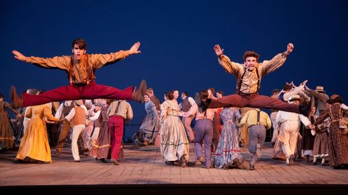 Two performers jumping in the air in the foreground. Other performers dressed in pioneer clothing are on the stage in the background. 