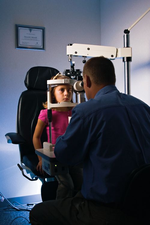 A young girl in a pink shirt sits in a tall black chair and rests her head on an eye examination device while the doctor examines her eyes.