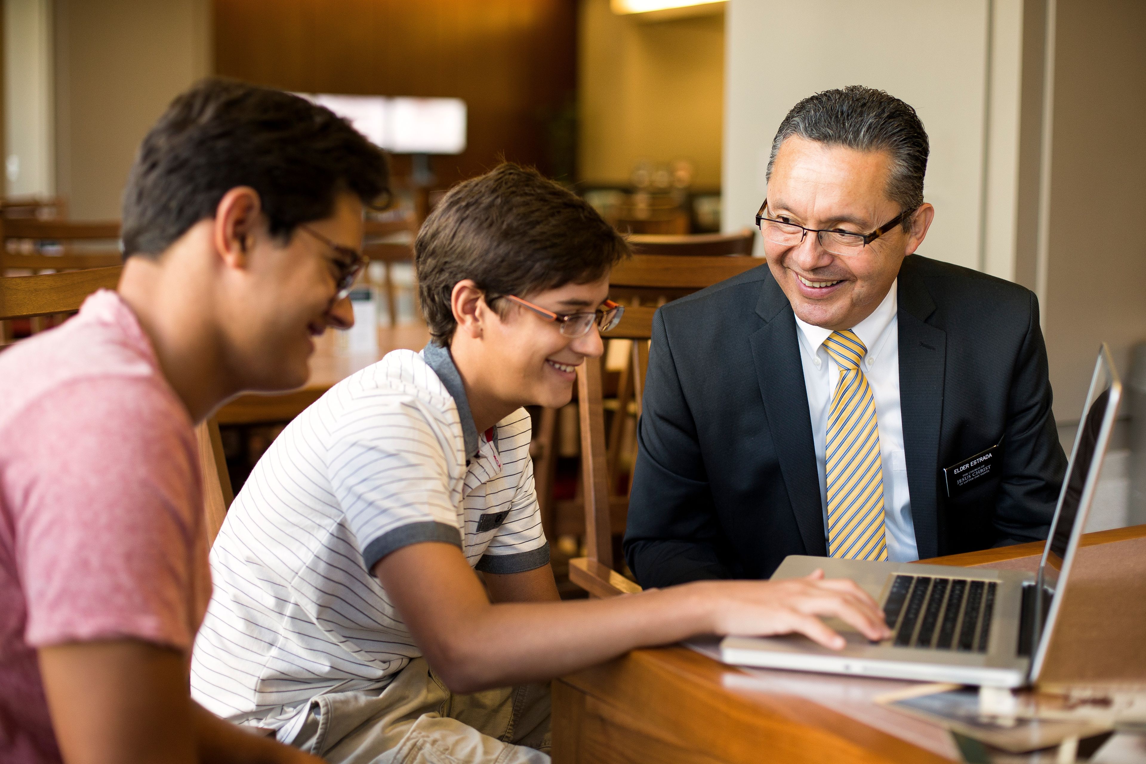 Two young men do family history on a laptop with a senior elder missionary.  
