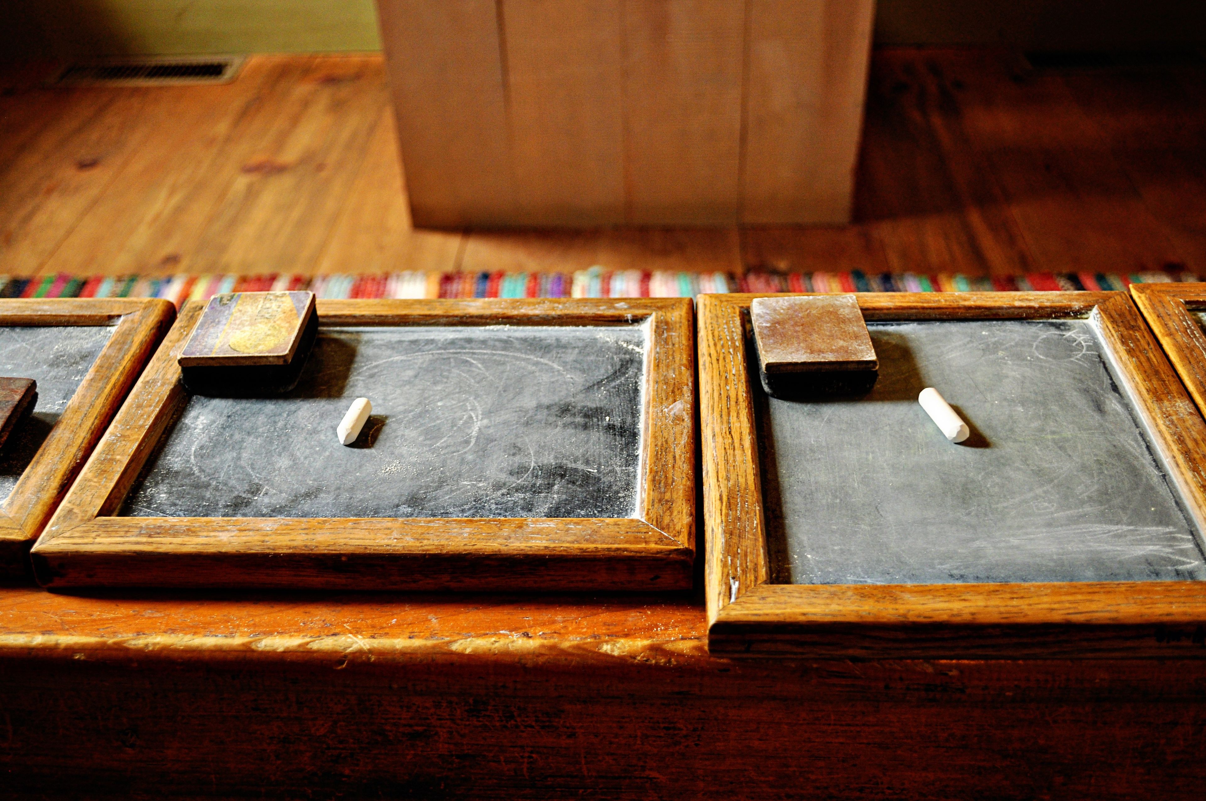Several small chalkboards with chalk and erasers are lined up on a bench in a schoolhouse.