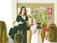 A woman leading music in Primary.  A girl is standing at her side holding a copy of the Book of Mormon.