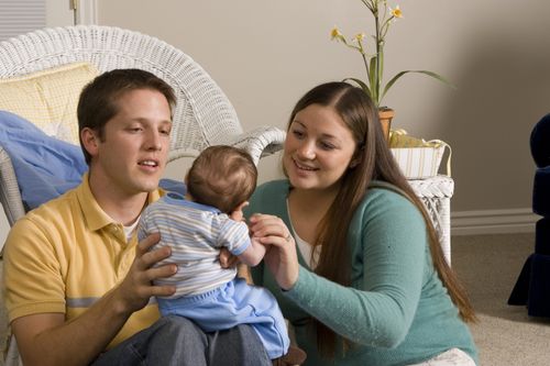 A photograph of a father and mother sitting on a living room floor and holding a baby boy.