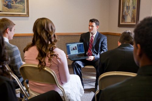 A male Sunday School teacher, in a suit and tie, sits at the front of a classroom full of young men and women and shows them a video from his laptop.