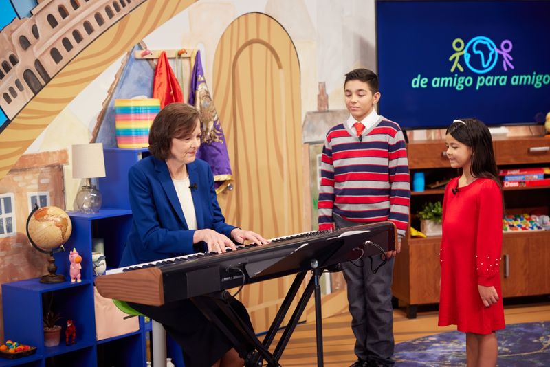 Sister Susan H. Porter sits at an electric keyboard on the set for the 2022 Friend to Friend broadcast while recording a segment. Rodrigo Filho and Mayte Pereira, the child hosts of the broadcast, listen to her play.