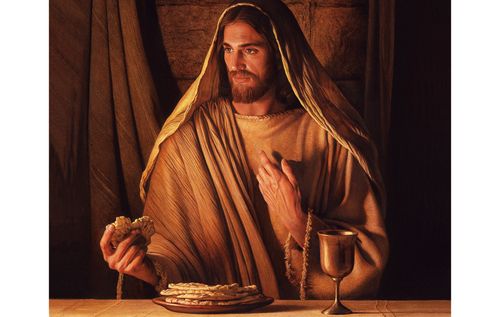 Jesus Christ seated at a table. In one hand He is holding a piece of bread. With the other He is gesturing towards Himself. There is a plate of bread on the table in front of Him and a goblet.