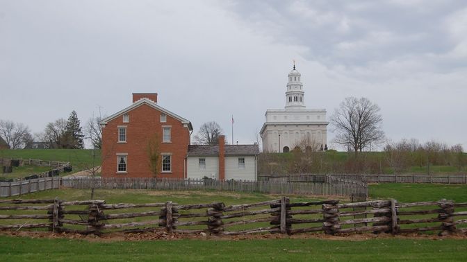 A fence in front of a two-story brick home with a single-story white clapboard addition. A white stone temple is atop the hill.