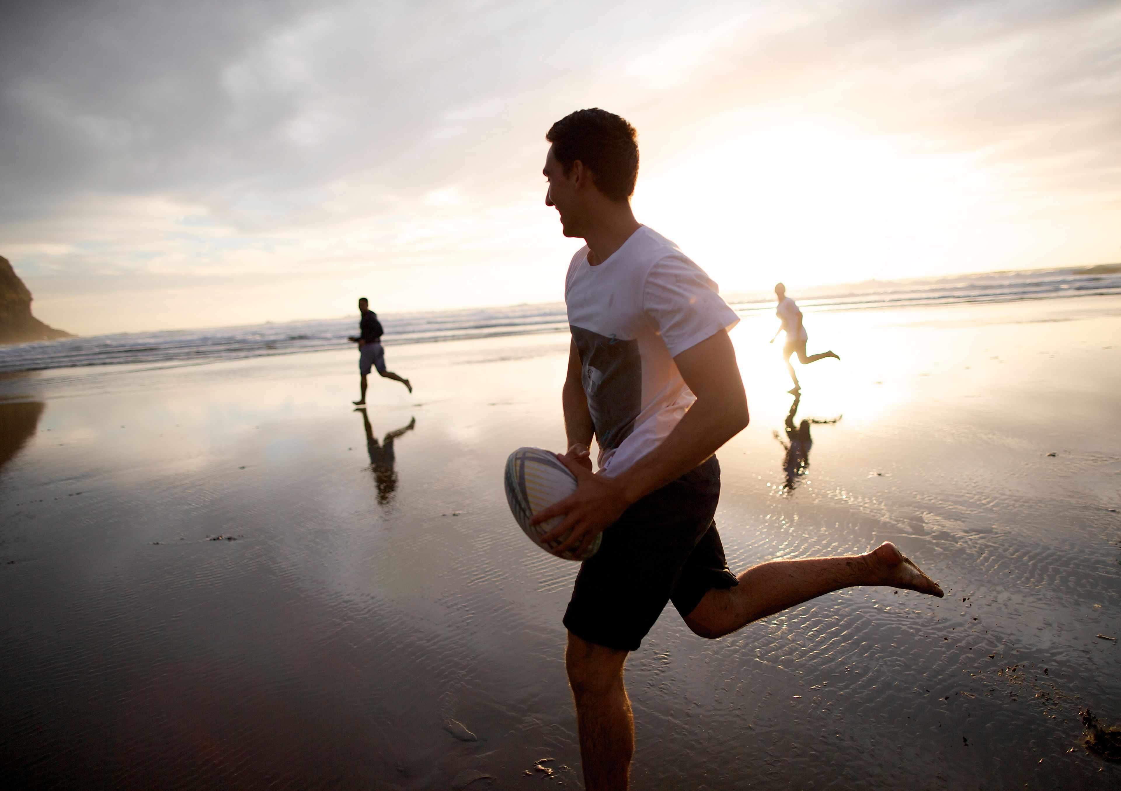 A young man playing rugby on the beach, running with the ball as the sun sets.  