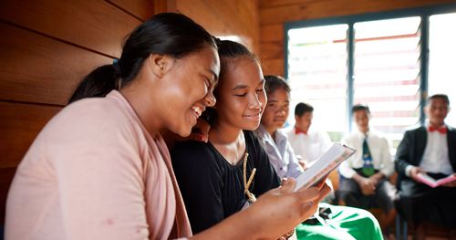 Various youth gather together. They appear to be in church in a Sunday School class. They have scriptures and smile. This is in Samoa.