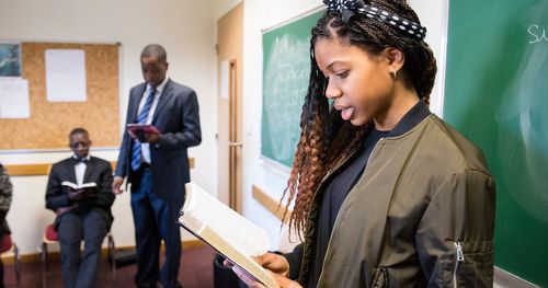 A group of teenagers sit in a classroom learning together while a young woman reads from the scriptures, the other youth follow along. The teacher can be seen standing to the side.