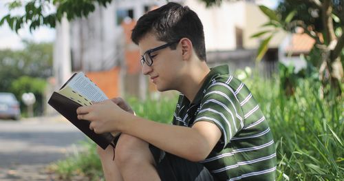 Young man wearing eyeglasses, sitting outside reading scriptures. (horiz)
