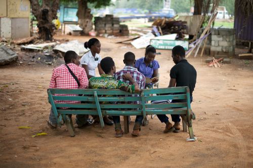 Two sister missionaries teaching a group of four teenage boys sitting on a green wooden bench in Ghana, Africa.